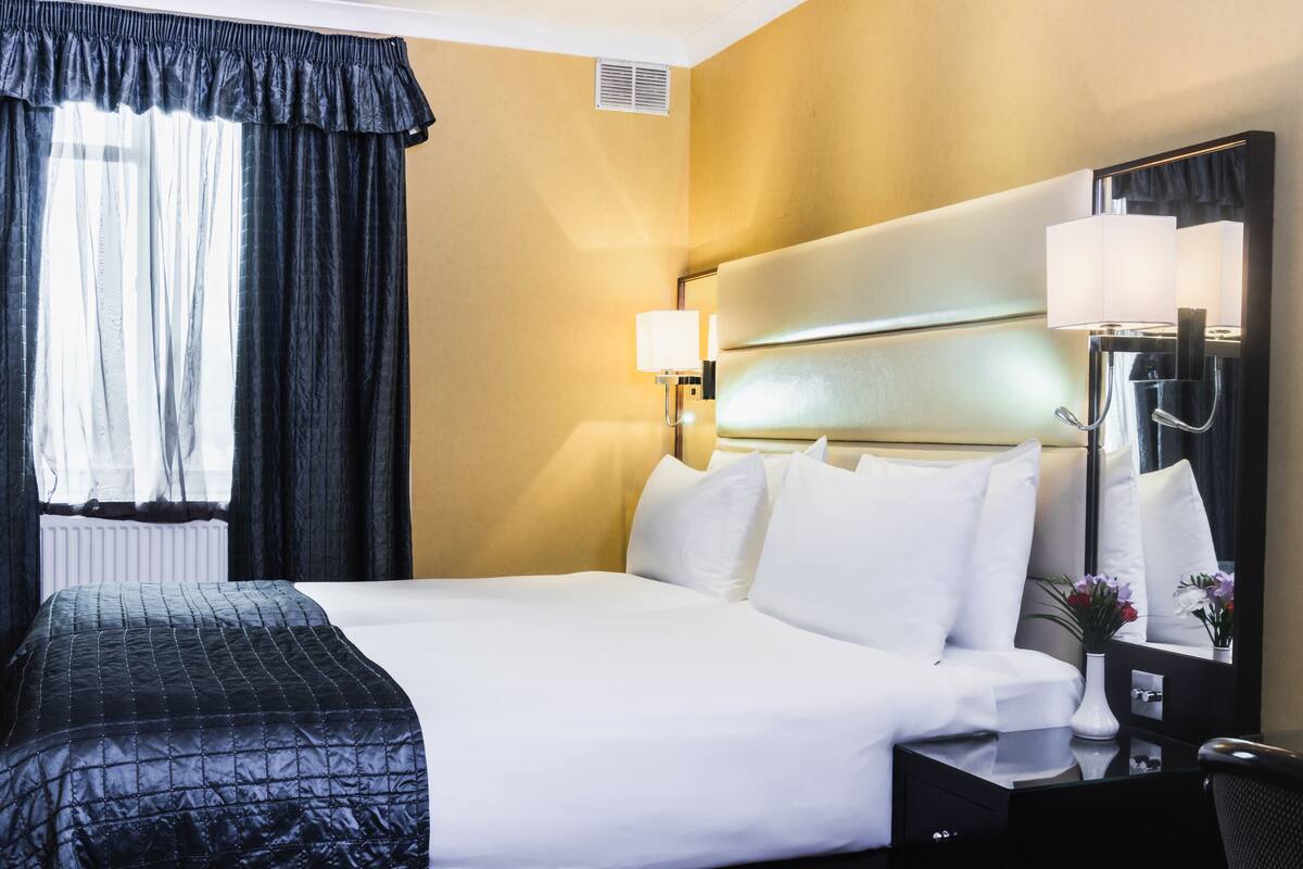 Une chambre d’hôtel avec un grand lit, une table de chevet avec une lampe, un miroir et une fenêtre avec des rideaux.