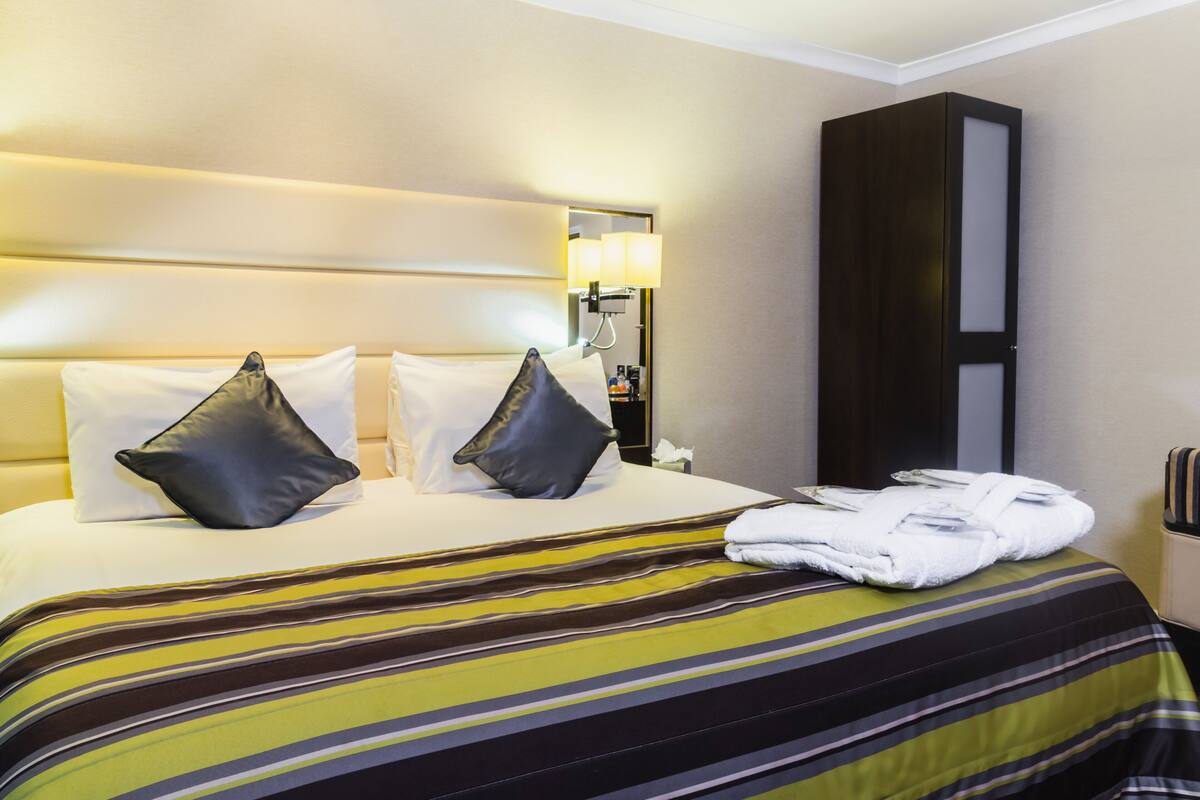 Une chambre d’hôtel avec un lit, des oreillers, une couvre-lit à rayures, une table de chevet avec une lampe et une armoire.