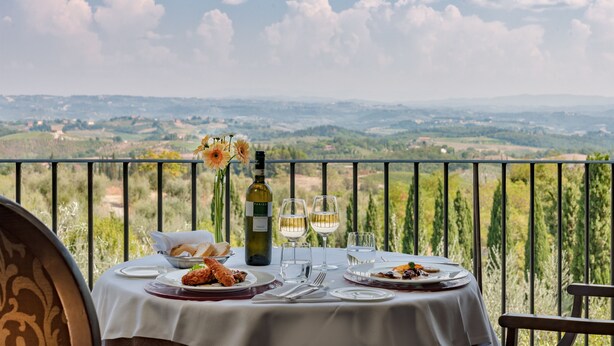 Une table dressée pour deux personnes, avec des assiettes remplies de nourriture, des verres à vin et une bouteille de vin, sur un balcon offrant une vue panoramique.