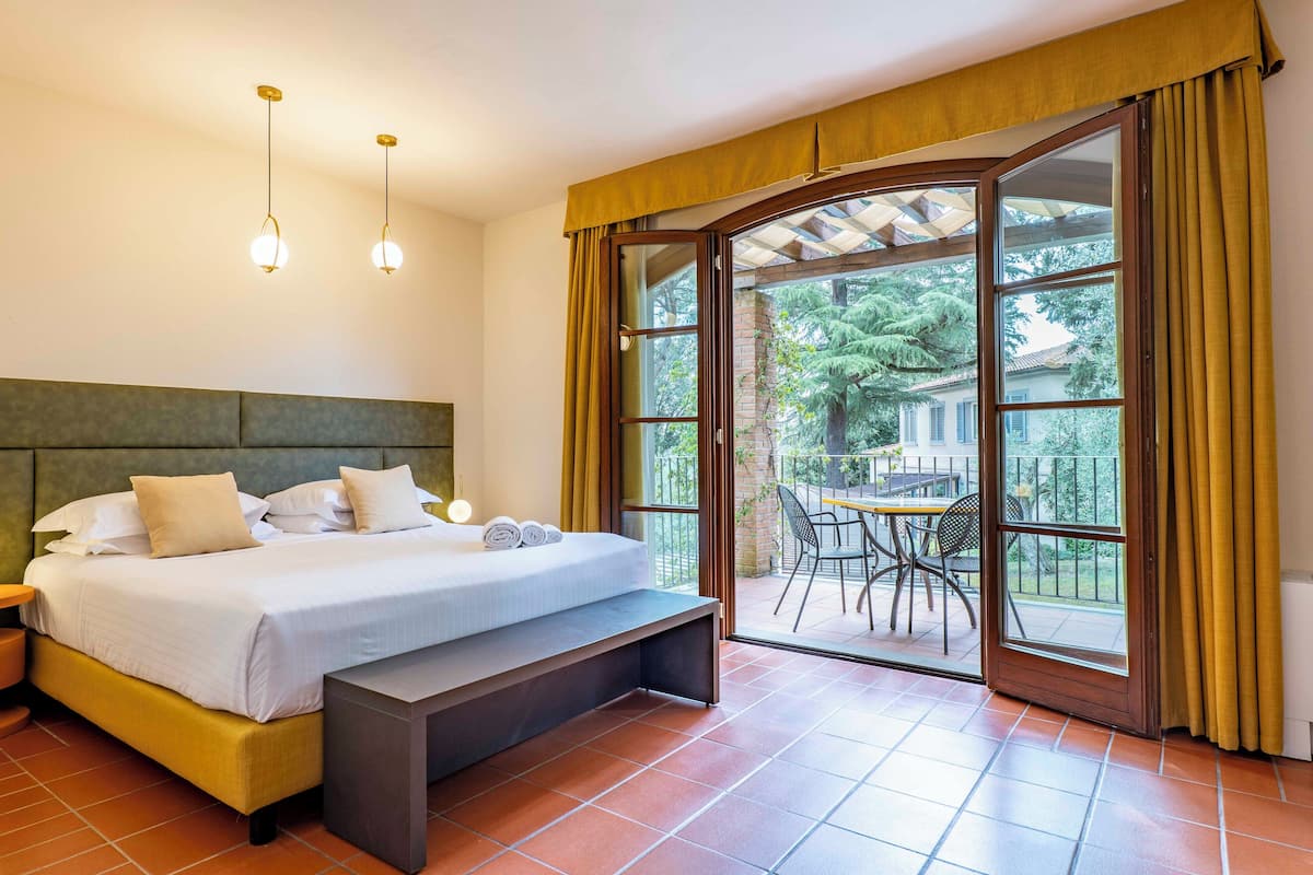 A bedroom with a bed, bench, bedside table, and a view of an outdoor patio with a table and chairs.