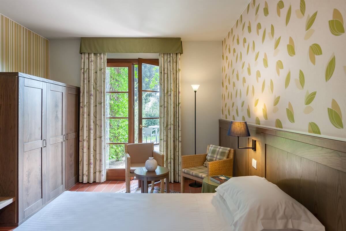 A hotel room with a bed, a chair, a table, a lamp, and a wardrobe.