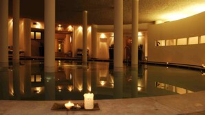 Un espace piscine intérieure paisible, avec une bougie posée sur un plateau, entouré de colonnes et éclairé d’une lumière douce.