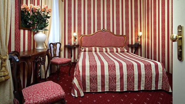 A hotel room with a striped wallpaper, a bed with a red and white striped bedspread, a wooden nightstand with a lamp, a chair, and a desk with a vase of flowers. - Hotel Centauro (Venice)