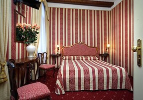 Quarto de hotel com papel de parede listrado, cama com colcha listrada vermelha e branca, mesa de cabeceira de madeira com abajur, uma cadeira e escrivaninha com um vaso de flores.