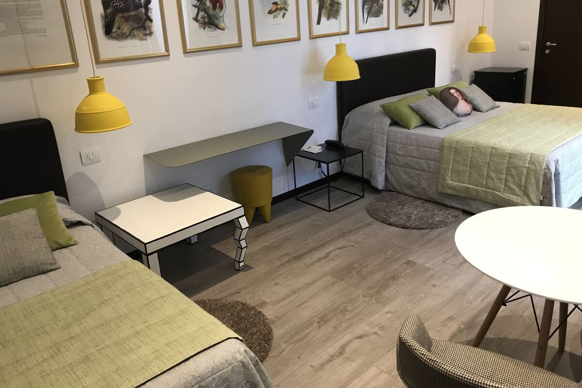 A modern hotel room with a bed, bedside table, desk, and chair. There are framed pictures on the wall and a yellow lamp.