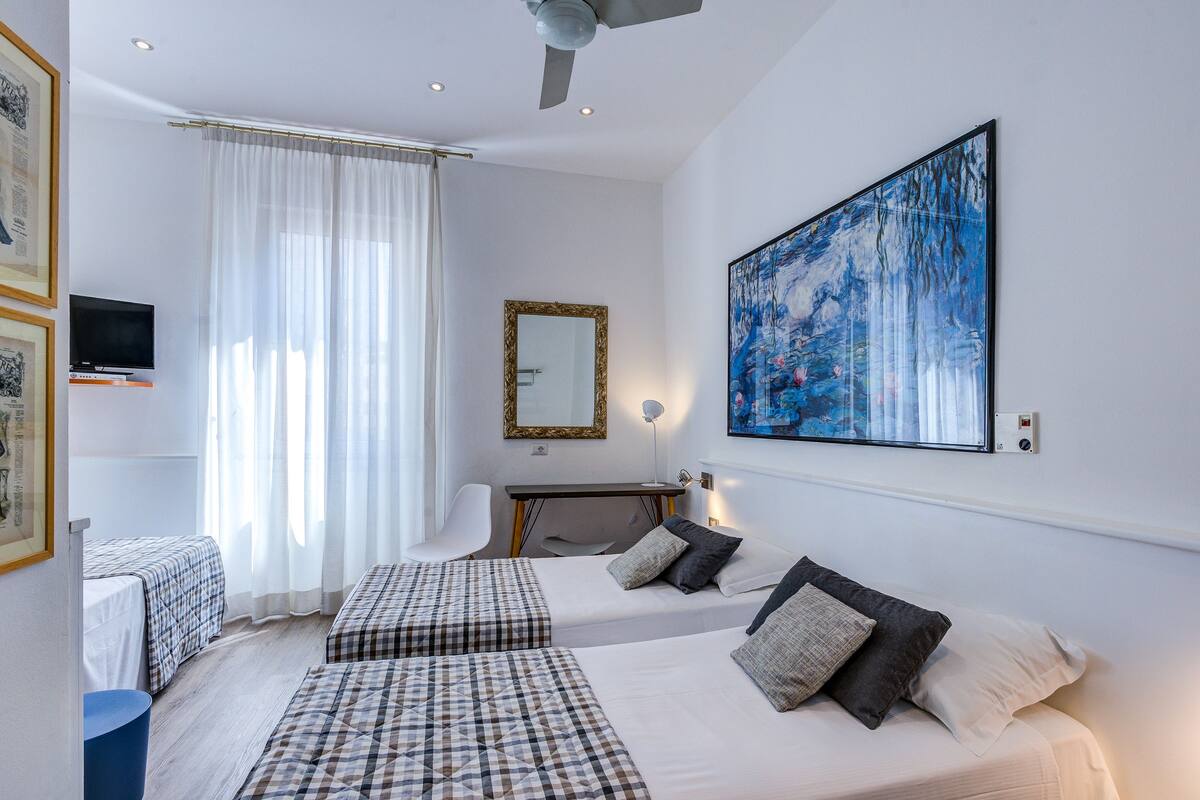 A hotel room with two beds, a desk, a chair, a television, a large painting, and a ceiling fan.