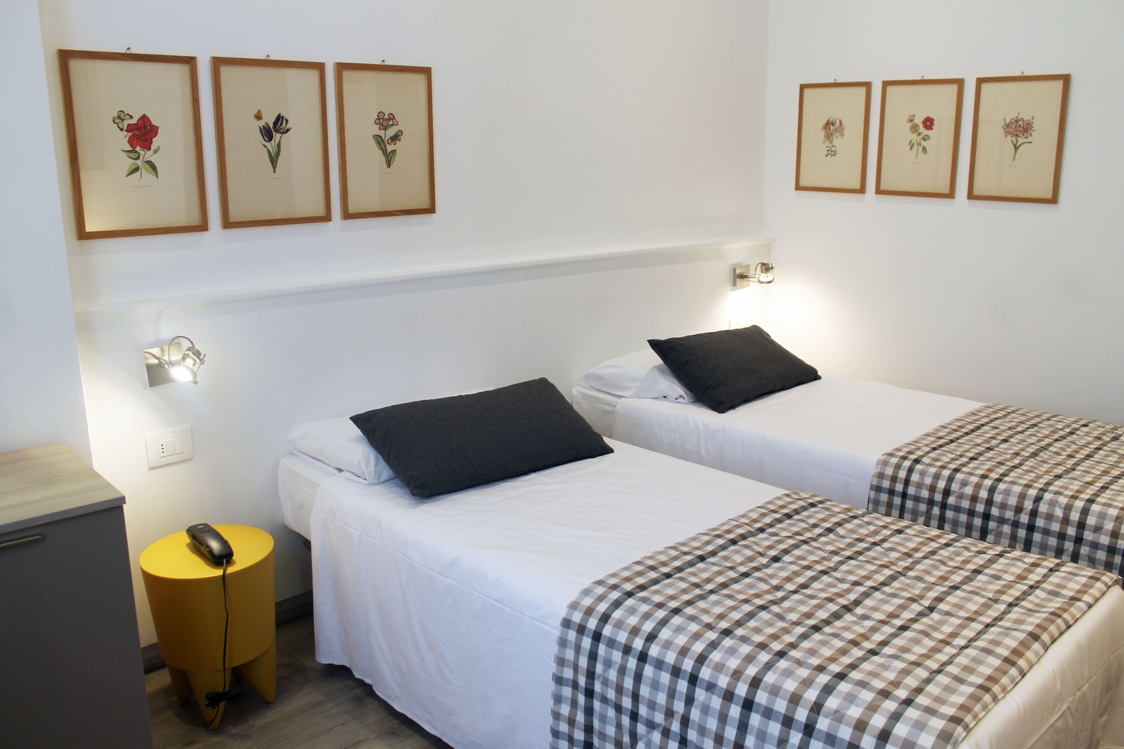 Een hotelkamer met twee eenpersoonsbedden, een nachtkastje met een lamp, een klein rond tafeltje met een telefoon, en bloemkunstwerken in een lijst aan de muur.