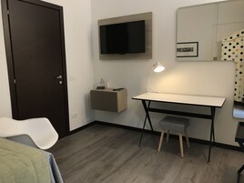 A compact room with a desk, a chair, a television mounted on the wall, a small stool, and a bed with a white cover.