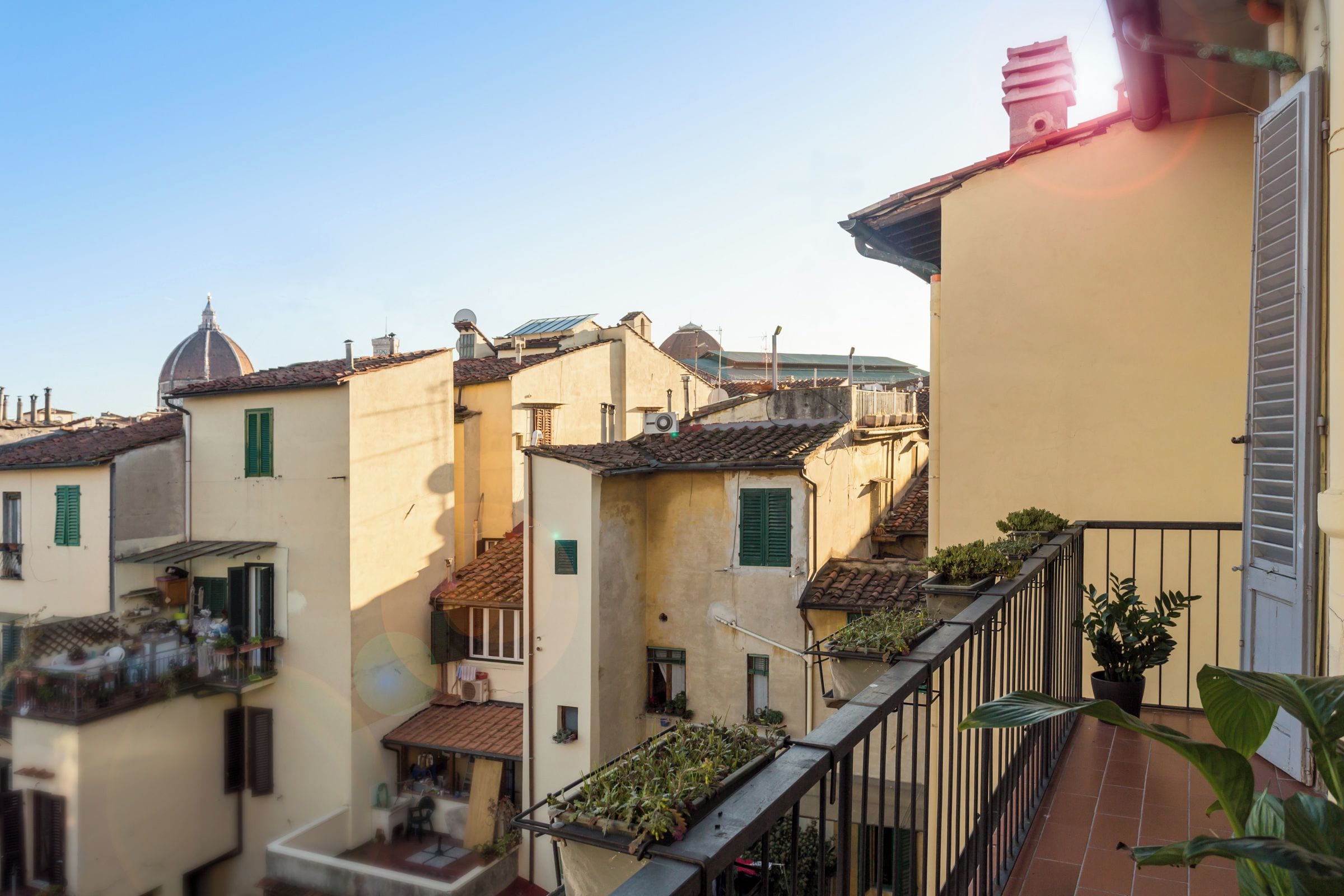 A narrow street with traditional buildings, a small balcony with potted plants, and a clear sky.