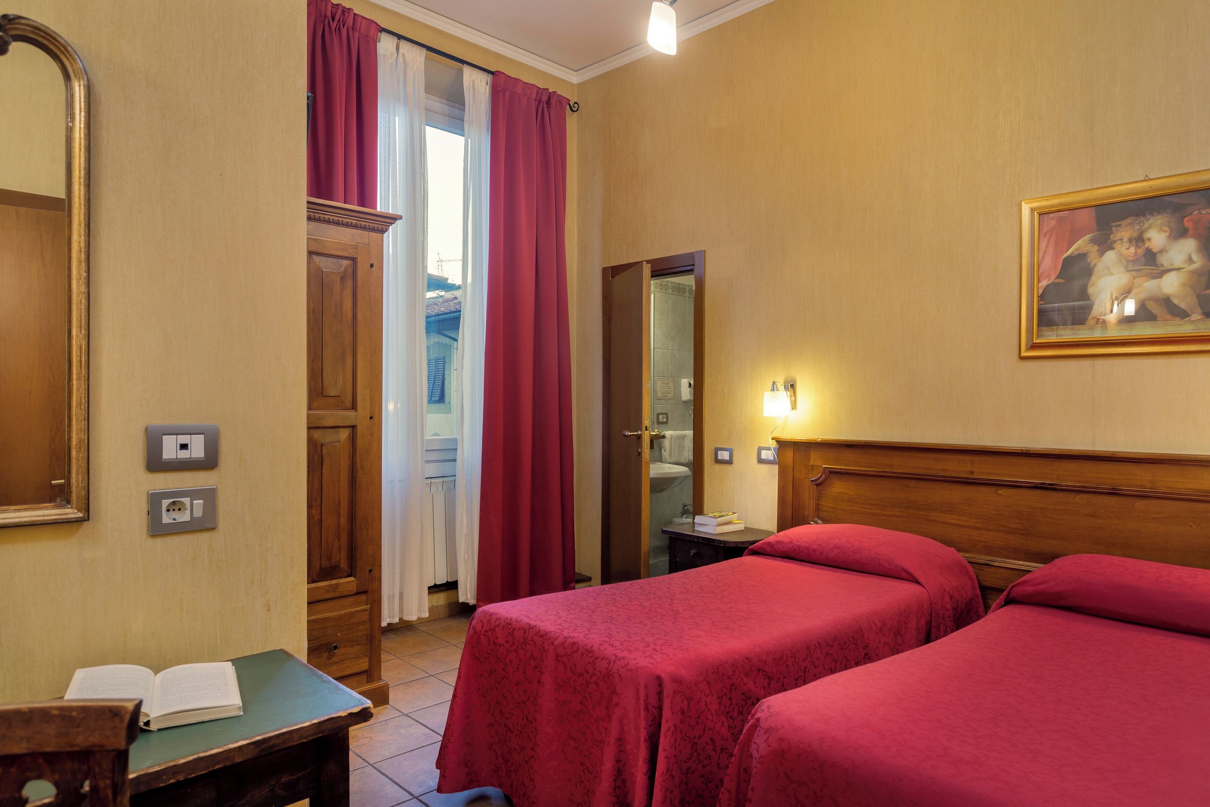 A hotel room with two single beds, a wooden desk with an open book, a mirror, a painting on the wall, and a bathroom visible through an open door.