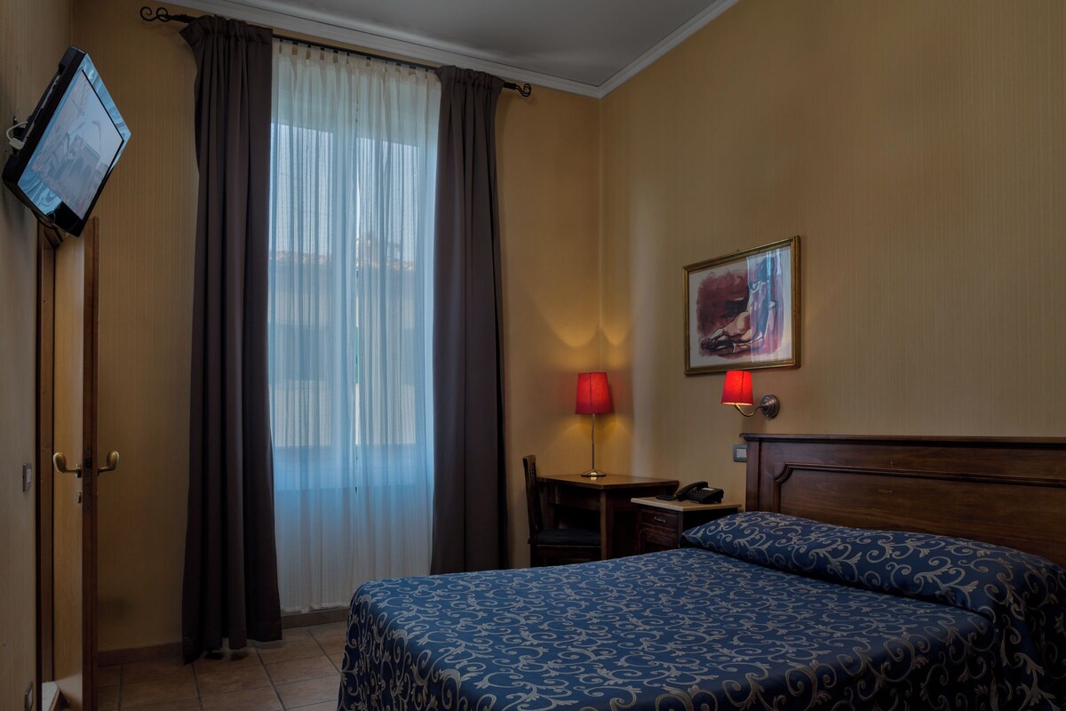 A hotel room with a bed, a desk, a chair, a TV, and a window with curtains.