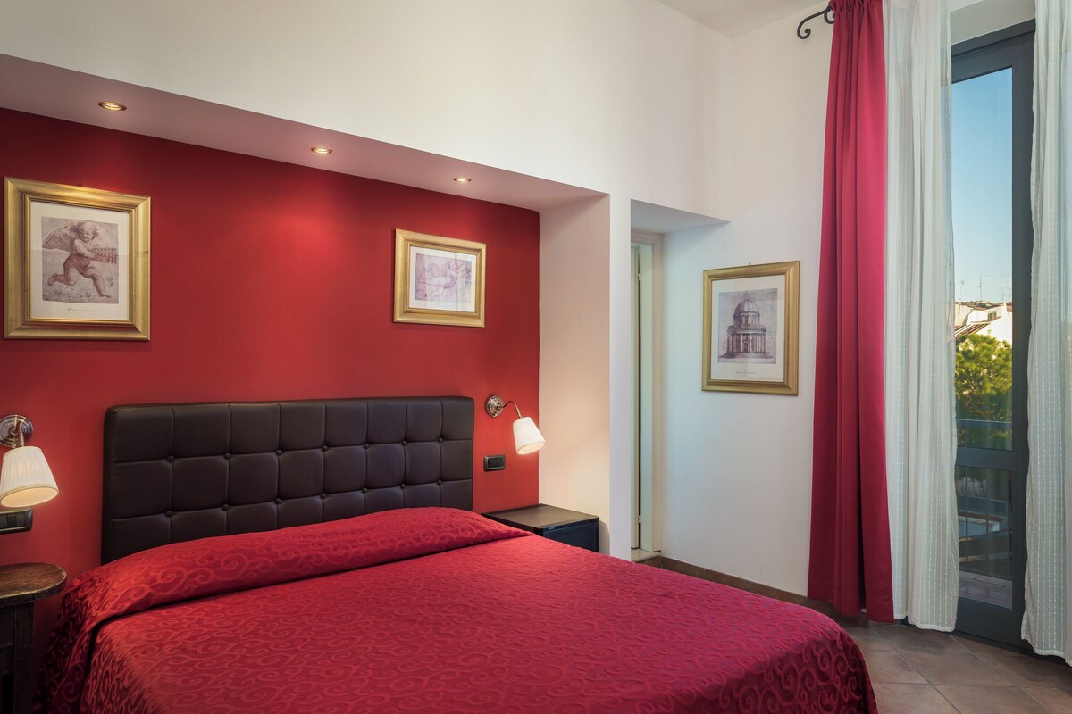 A hotel room with a red wall, a large bed, two framed pictures, a nightstand with a lamp, and a view through a window with curtains.