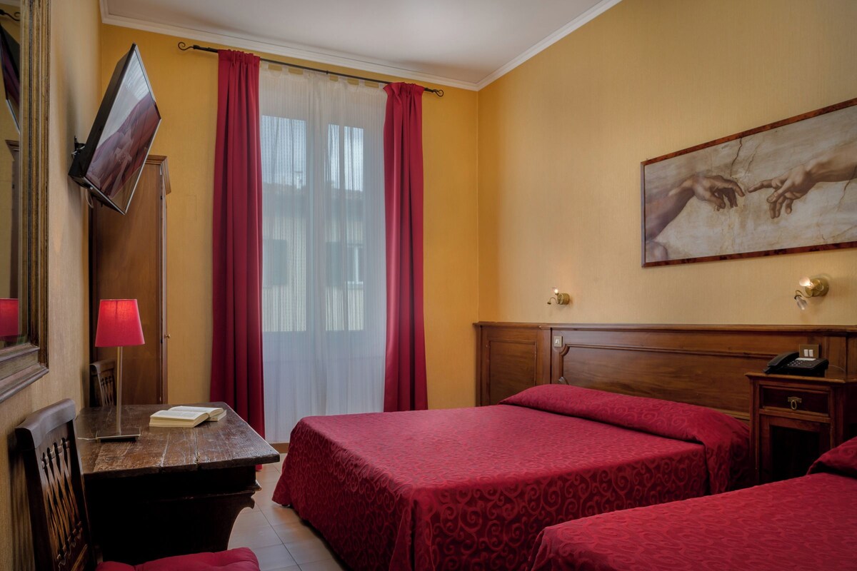 A hotel room with two beds, a desk with a lamp, a TV mounted on the wall, and a painting above the bed.