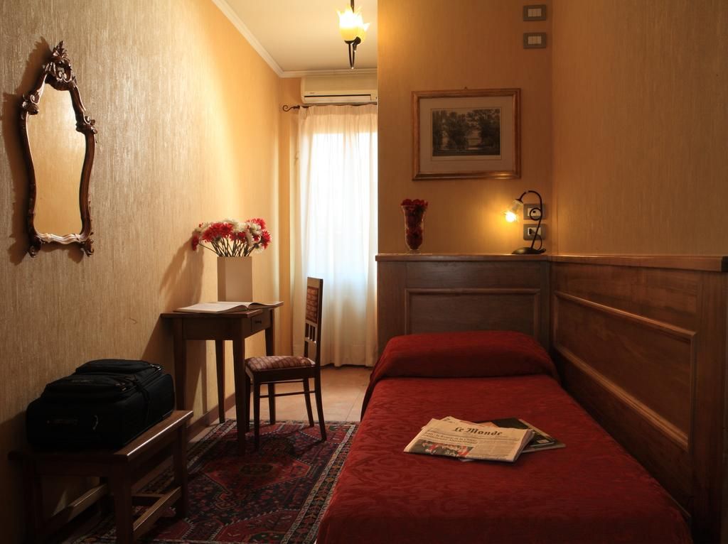 Une chambre d’hôtel avec un lit, un bureau, une chaise, un miroir, un vase avec des fleurs et un tableau encadré au mur.