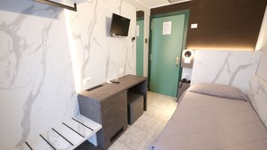 Quarto de hotel com cama, escrivaninha e cadeira, televisão na parede e porta verde.