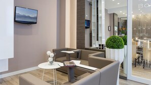 Uma área de espera moderna com televisão de tela plana na parede, um espaço de estar confortável com sofás e uma mesa pequena, e uma grande planta em vaso.