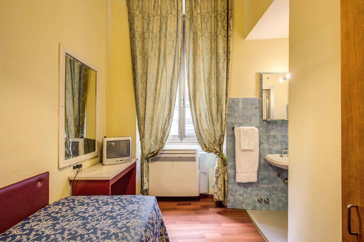 A small, compact hotel room with a bed, a desk with a television, a window with curtains, a bathroom with a sink and mirror, and a tiled floor.