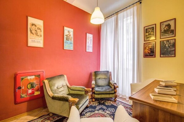 Um quarto com uma parede vermelha, duas poltronas, uma mesa de madeira com livros, um tapete estampado e um lustre.