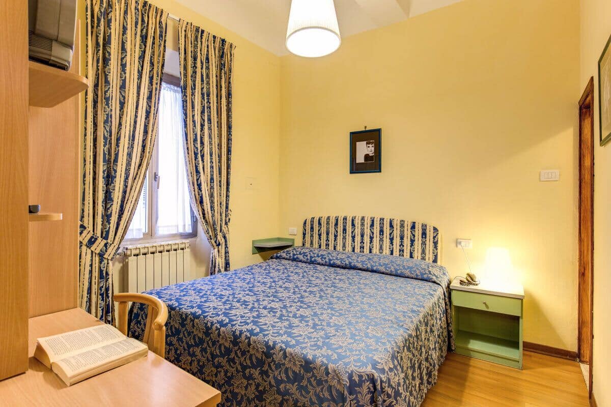 A bedroom with a bed, a bedside table, a chair, a desk, and a window with curtains.
