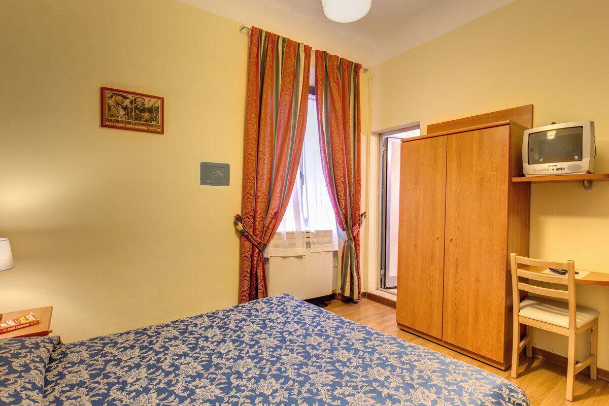 A bedroom with a bed, a wooden wardrobe, a television on a shelf, and a window with curtains.