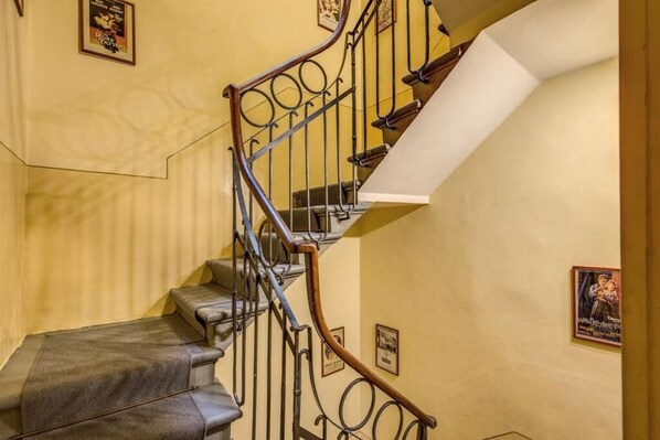 Una escalera con barandilla de madera y balaustradas de metal, que conduce a un rellano con pared blanca y cuadros enmarcados.