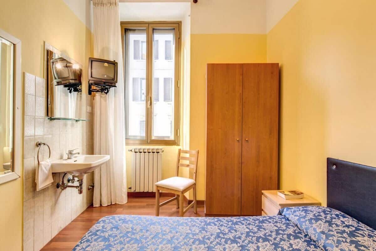 A bedroom with a bed, a wooden wardrobe, a small bedside table with a book, a chair, a window with curtains, a sink, and a mirror.