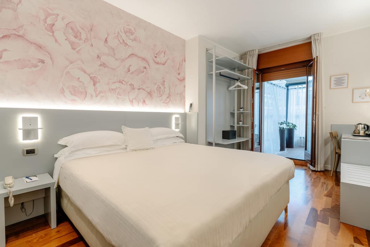 A hotel room with a large bed, bedside table with a phone, a wardrobe, and a desk with a kettle.