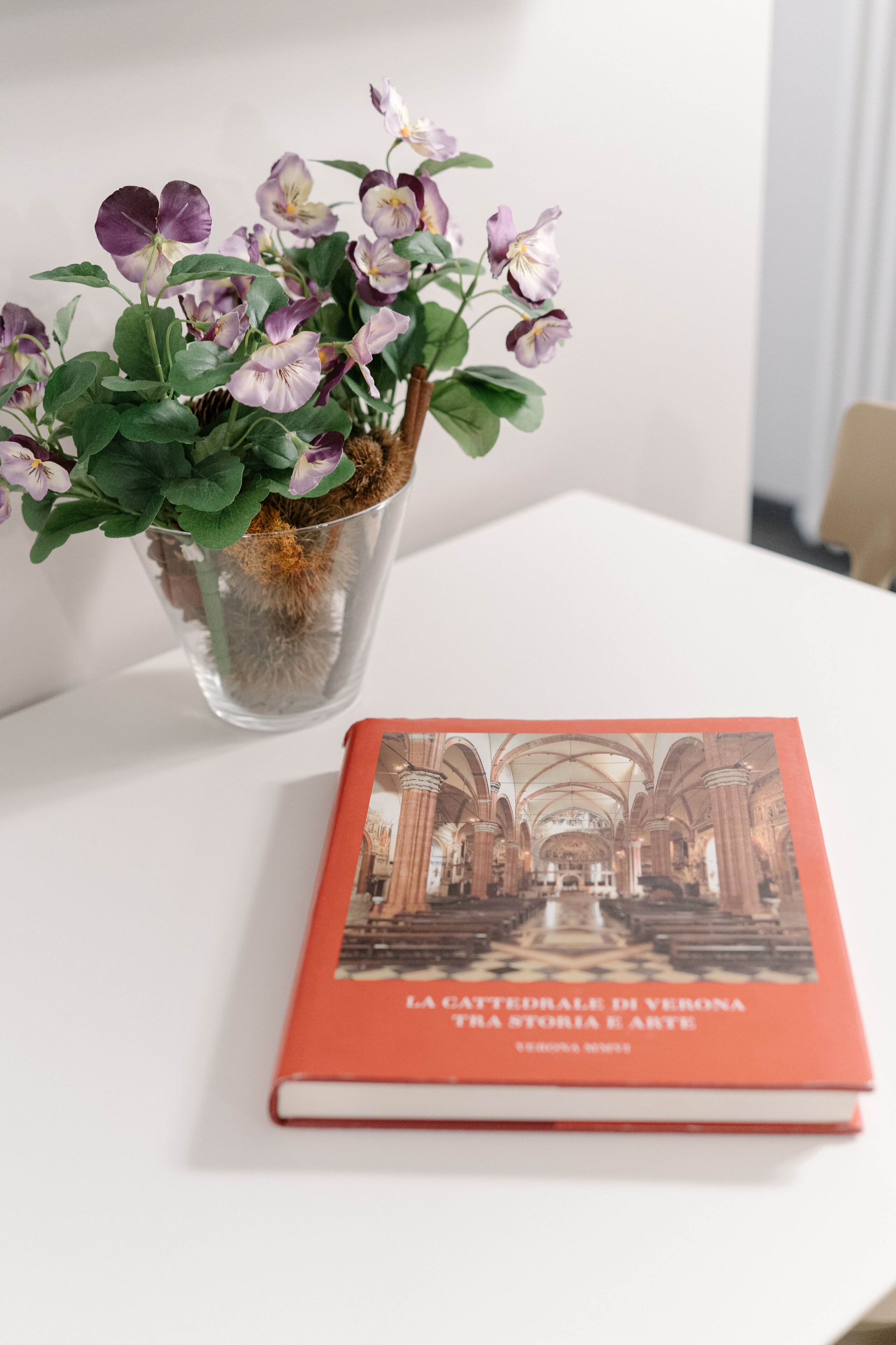 A book titled "La Gatedriale di Verona" next to a clear glass vase with purple flowers.