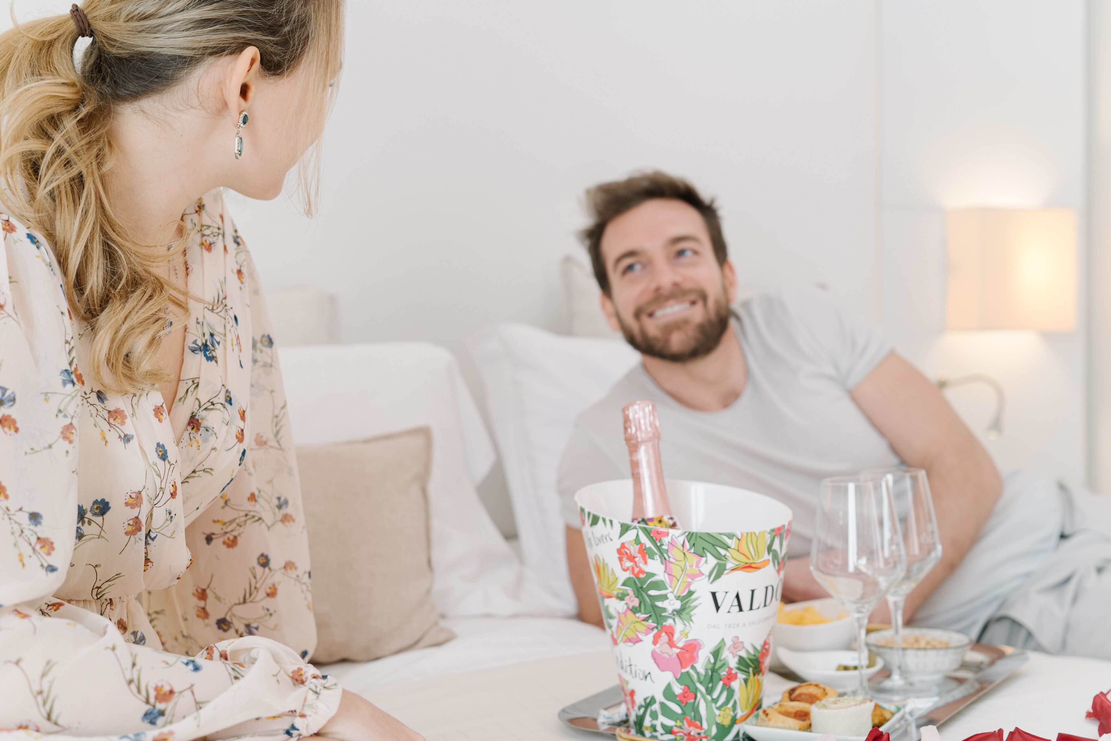 Una habitación con una cama, una mesita de noche con una botella de champán, un bol de comida y una copa.