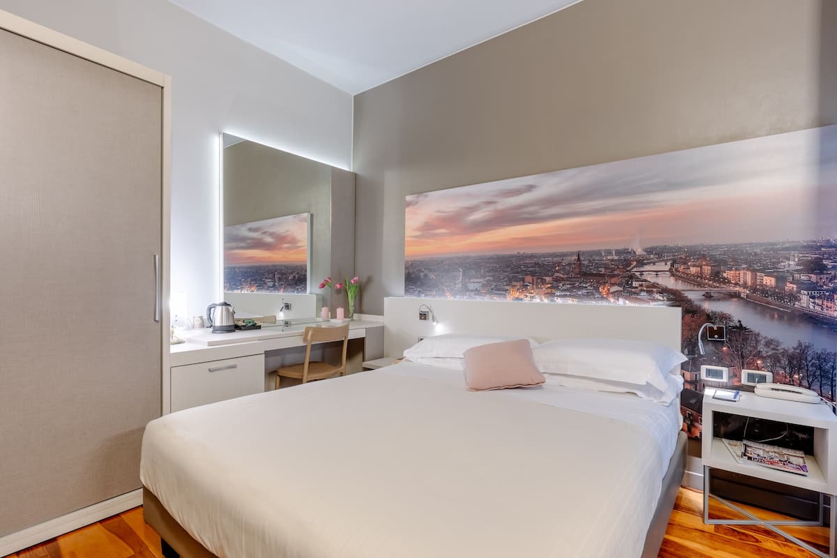 A modern hotel room with a large bed, a desk, a chair, and a view of a cityscape at sunset.
