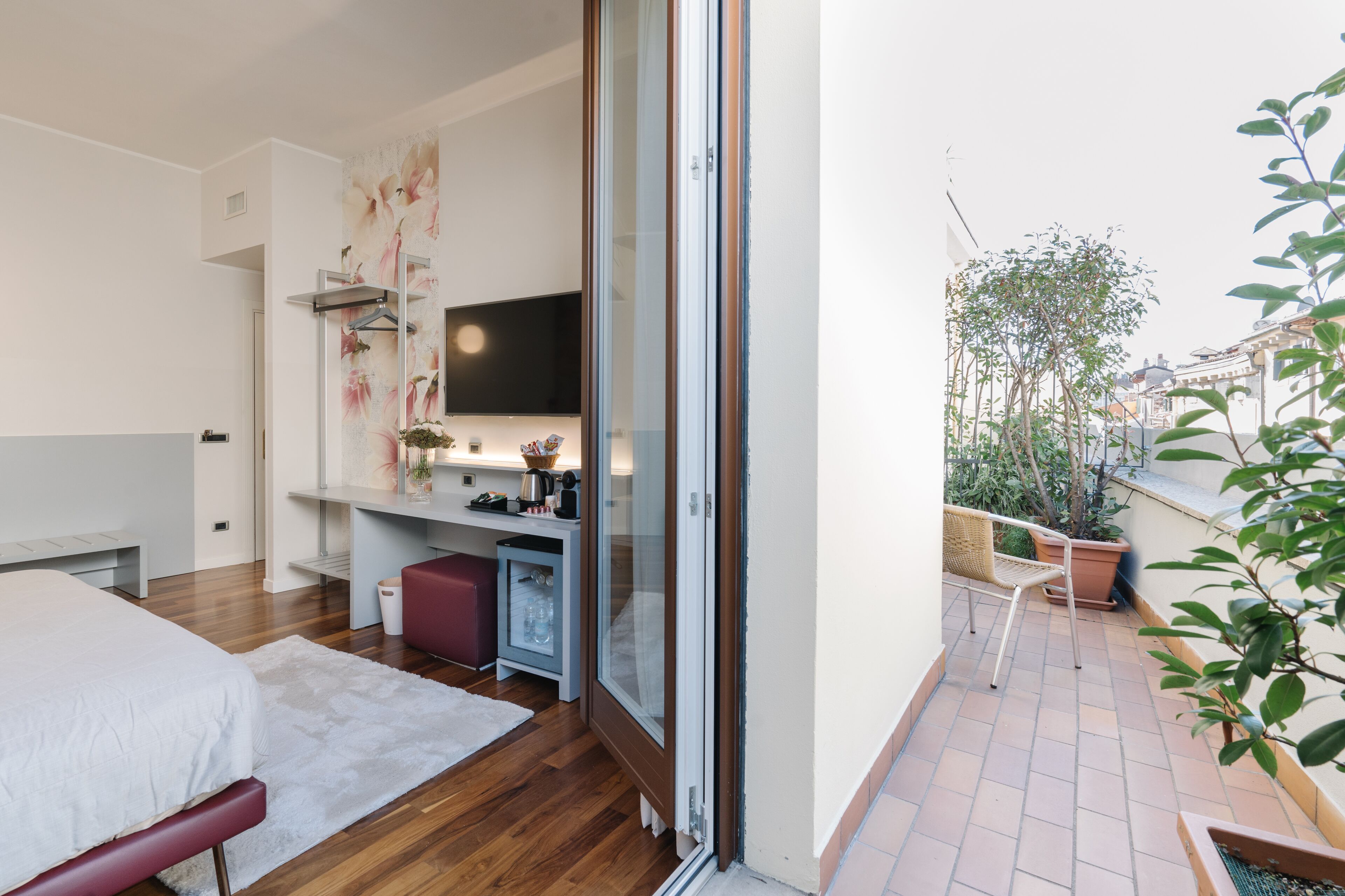 Un dormitorio con cama, escritorio, televisión y puerta corrediza de vidrio que da a un balcón con plantas y vistas a edificios.