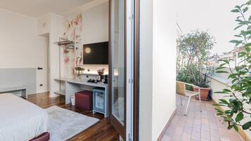 Un dormitorio con cama, escritorio, televisión y puerta corrediza de vidrio que da a un balcón con plantas y vistas a edificios.