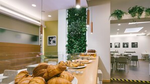 Un bufé de desayuno con cruasanes y bollería, un jardín vertical verde y un interior moderno con mesas y sillas.