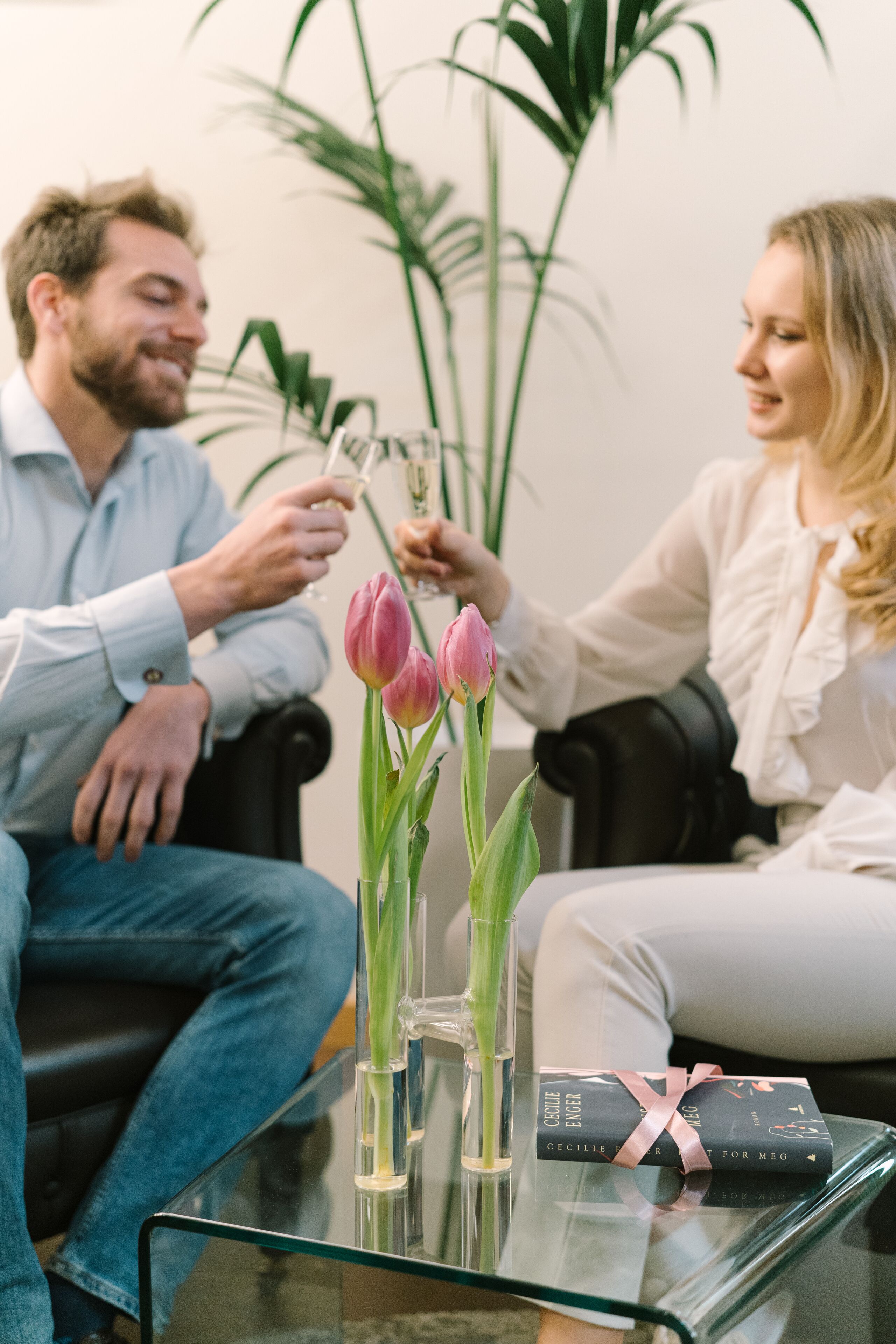 Ein Mann und eine Frau sitzen auf einer Couch, vor ihnen ein Glastisch, eine Vase mit Tulpen und ein verpacktes Geschenk.