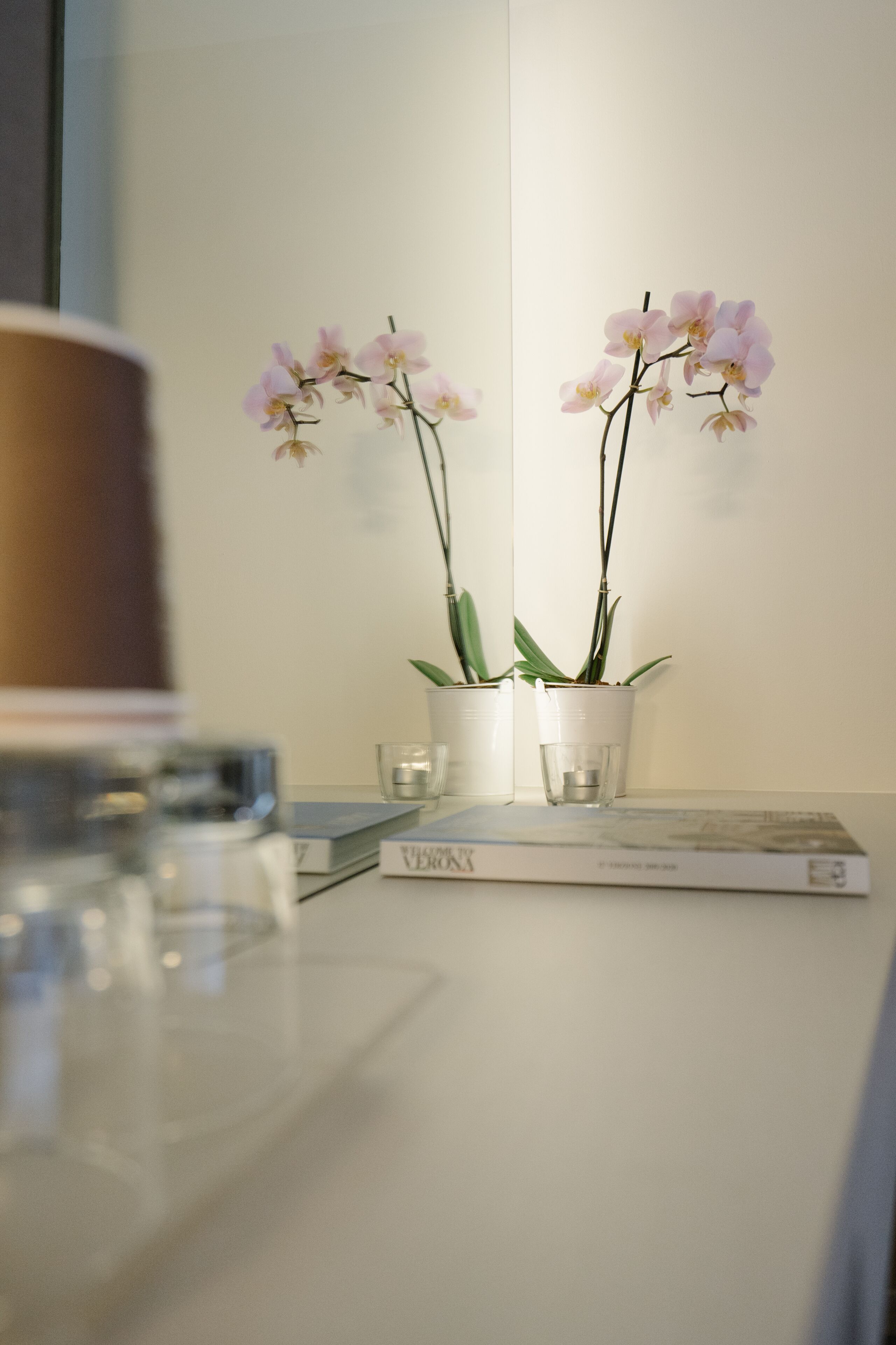 Uma mesa branca com duas orquídeas em vasos, um copo e um livro intitulado 