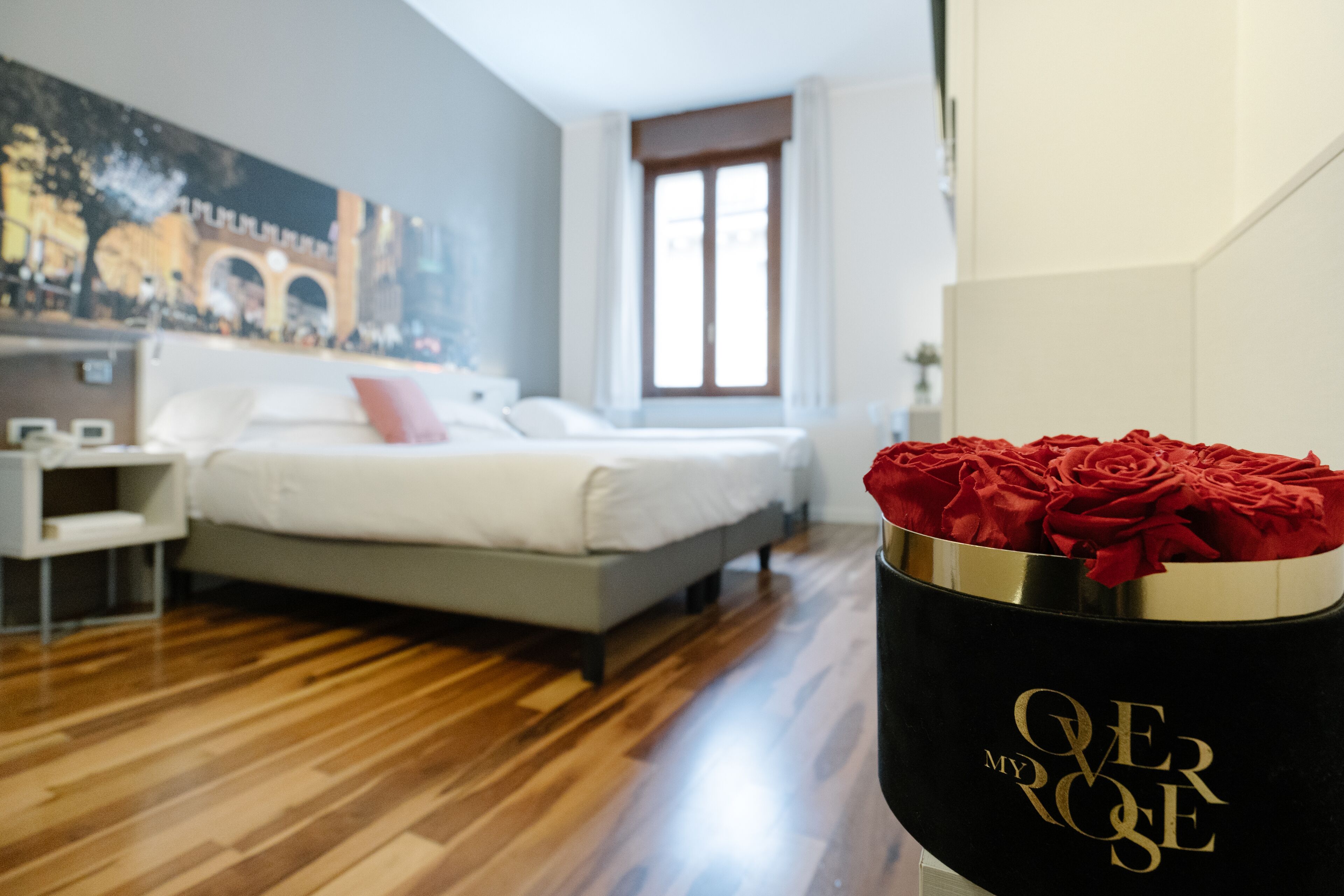 A modern bedroom with a bed, a nightstand, a vase with red roses, and a mural on the wall.