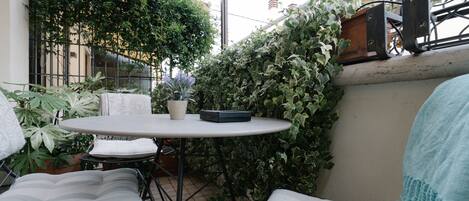 Un patio al aire libre con una mesa redonda y dos sillas, rodeado de abundante follaje y plantas en macetas.