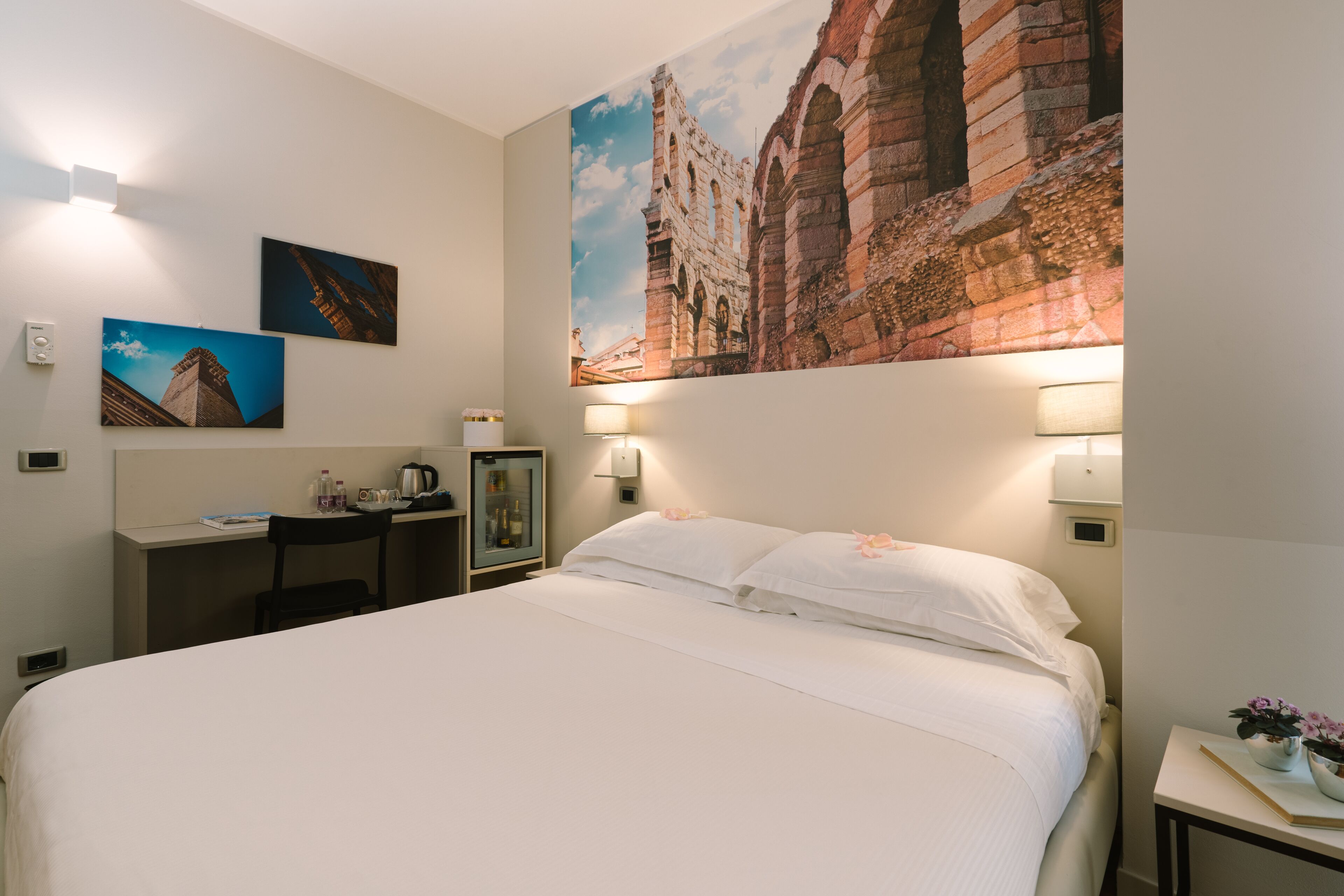A hotel room with a large bed, a desk with a chair, a small table with a lamp, and a wall-mounted TV.