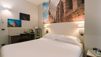 Quarto de hotel com uma cama grande, uma escrivaninha com cadeira, uma mesinha de cabeceira com abajur e uma TV fixada na parede.