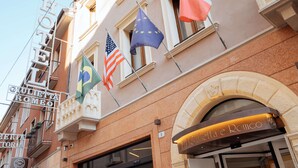 Fachada de hotel con banderas, incluyendo la estadounidense y la de la Unión Europea, y un letrero que dice 'Hotel Giulietta e Romeo'.