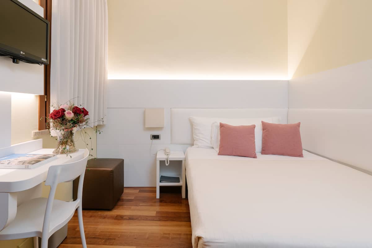 A hotel room with a bed, bedside table, chair, desk, and a small table with a vase of flowers.