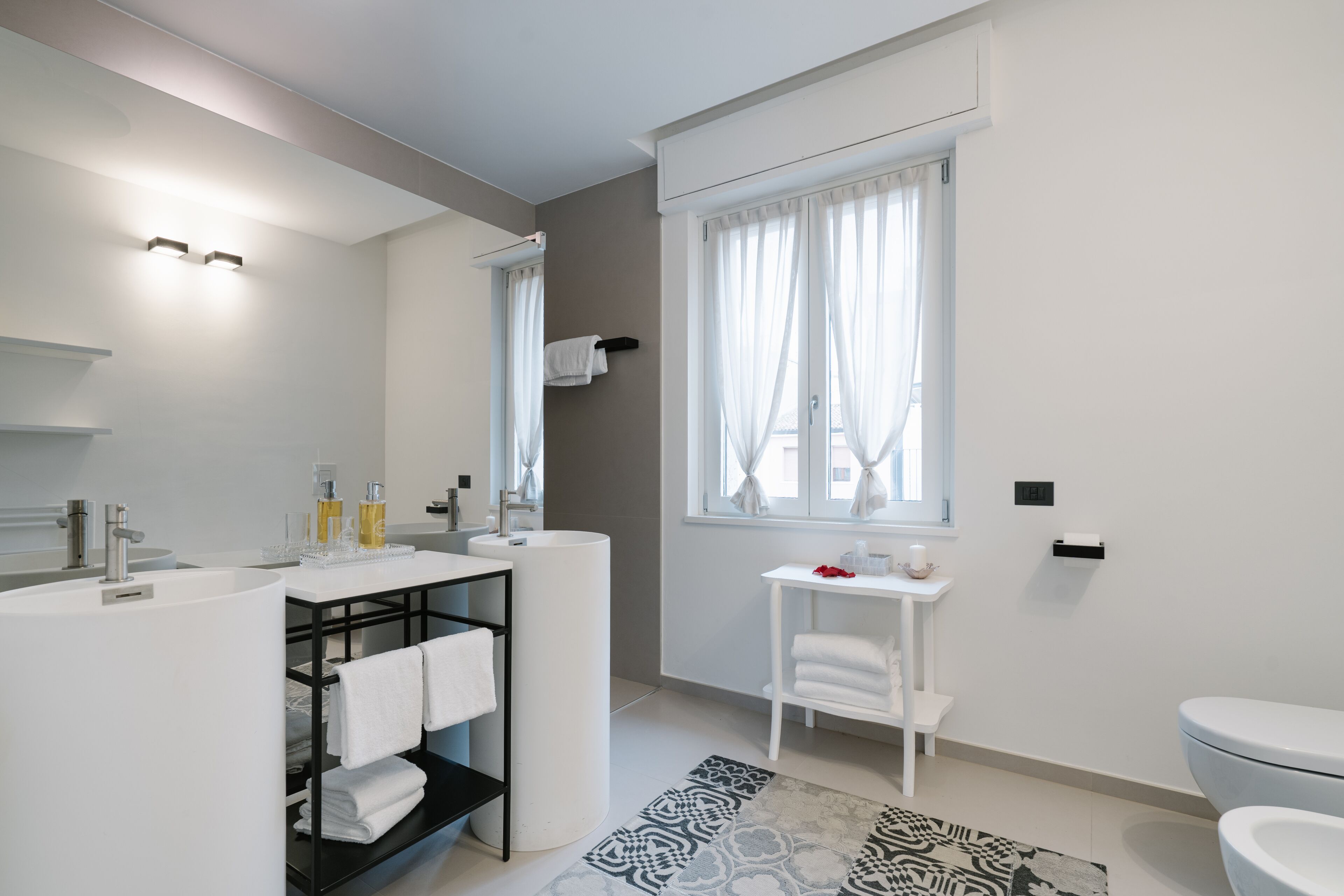 A modern bathroom with a white sink, vanity, and toilet. There is a window with curtains, a towel rack, and a patterned rug.