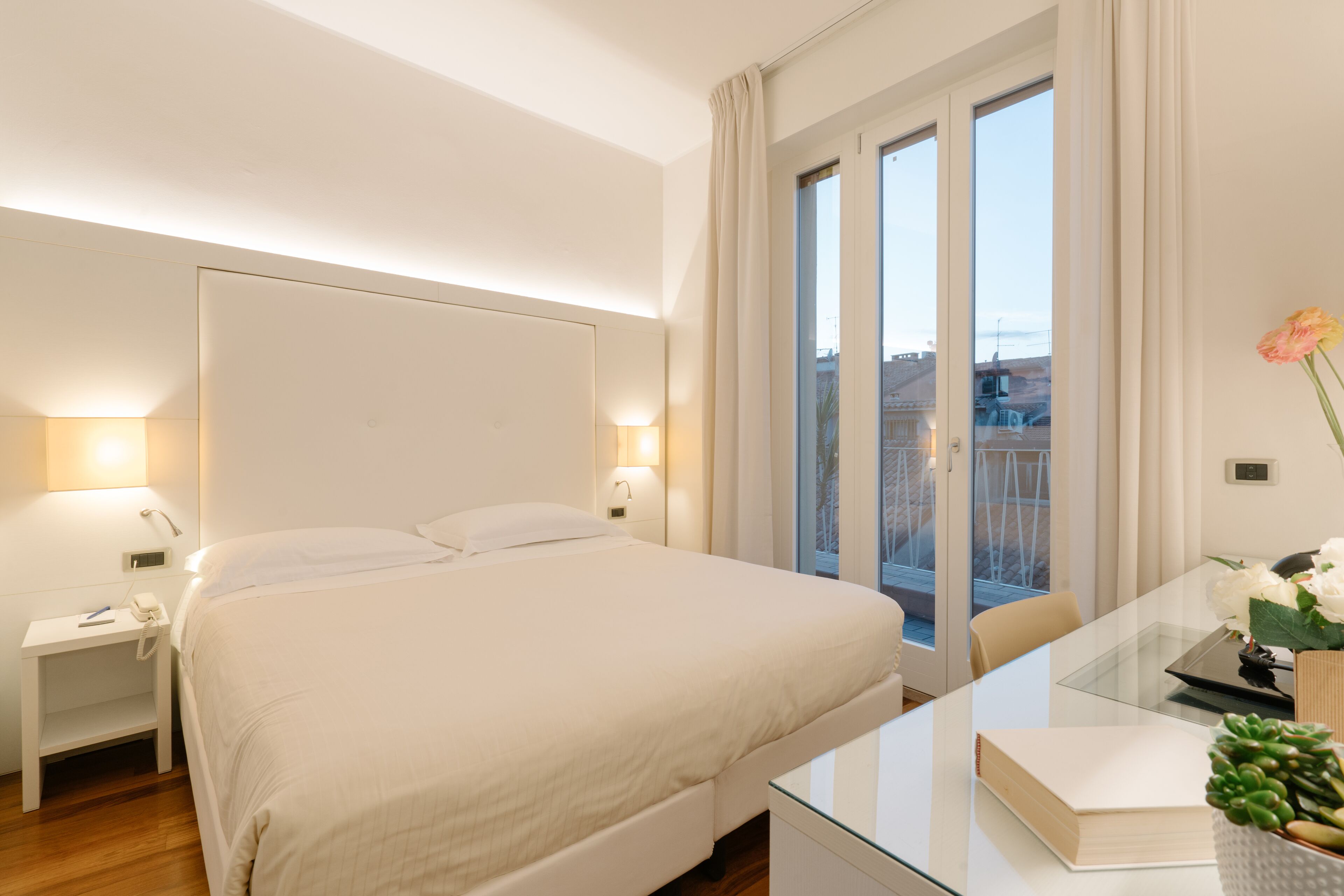 Ein Hotelzimmer mit einem großen Bett, Nachttischlampen, einem Glastisch und Blick auf die Stadt durch das Fenster.