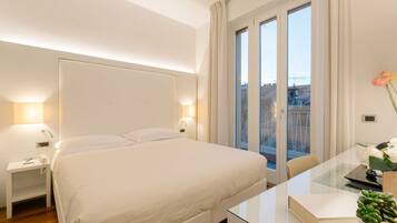 Ein Hotelzimmer mit einem großen Bett, Nachttischlampen, einem Glastisch und Blick auf die Stadt durch das Fenster.