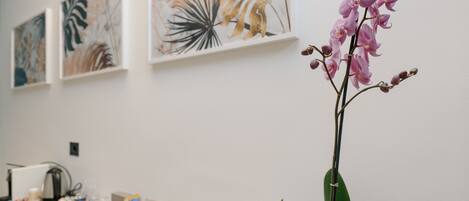 Una habitación con pared blanca, un jarrón con orquídeas rosadas, una cafetera y una cesta con libros.