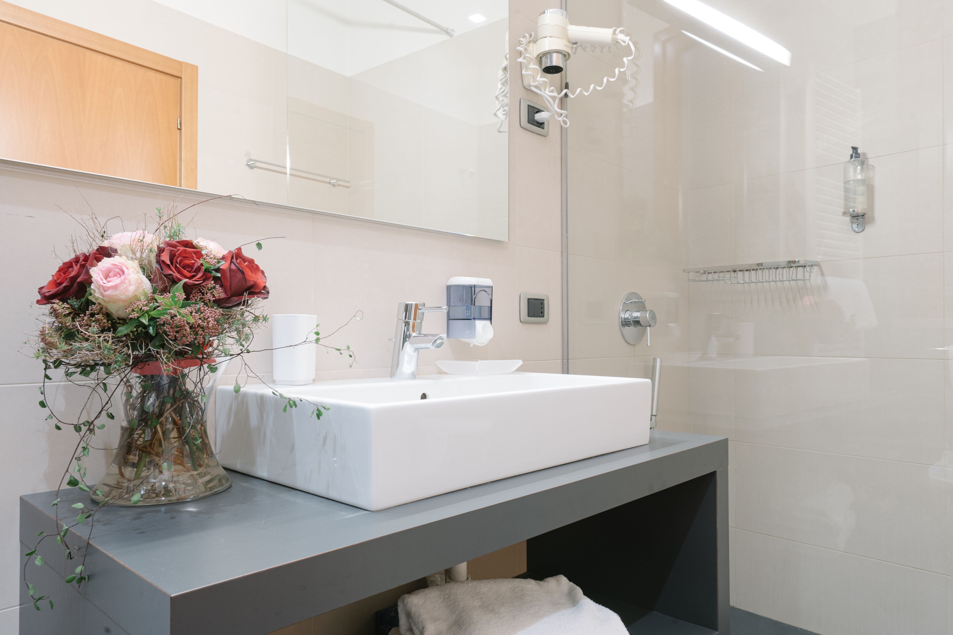 A bathroom with a white sink, a mirror, a vase with flowers, and a towel rack.