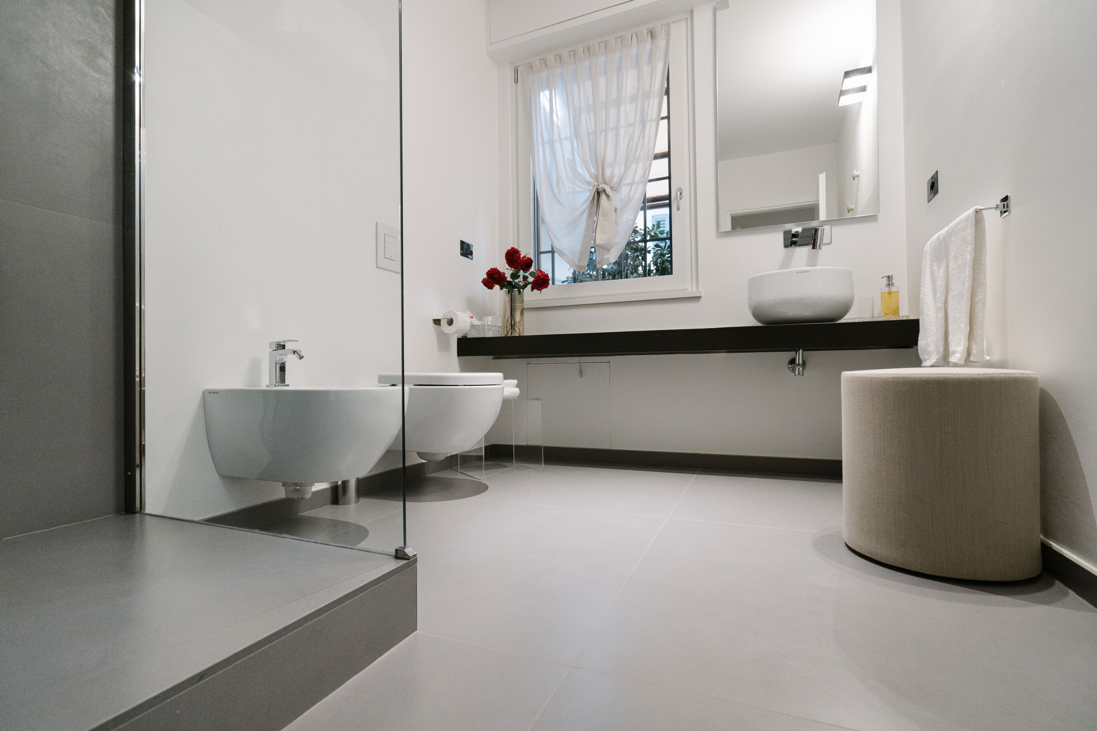 Une salle de bain moderne avec une grande baignoire, un lavabo séparé et des toilettes. On y trouve une fenêtre avec des rideaux légers, un miroir au-dessus du lavabo et un porte-serviettes fixé au mur.