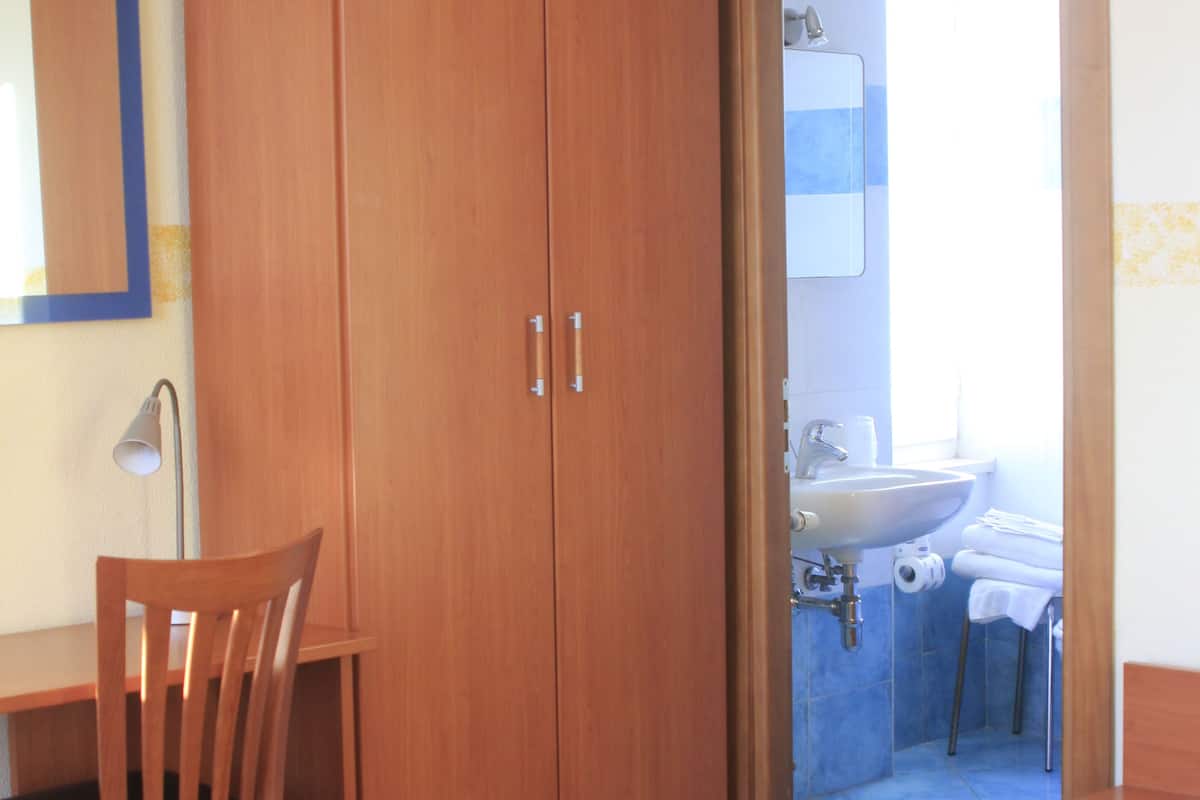 Une pièce comprenant une armoire en bois, un bureau avec une chaise, un lit recouvert d’une courtepointe, et une salle de bain visible à travers une porte ouverte.