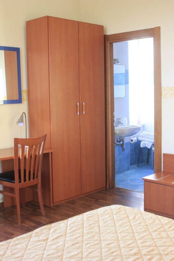 Una habitación con un armario de madera, un escritorio con una silla, una cama con cubrecama acolchado y un baño visible a través de una puerta abierta.