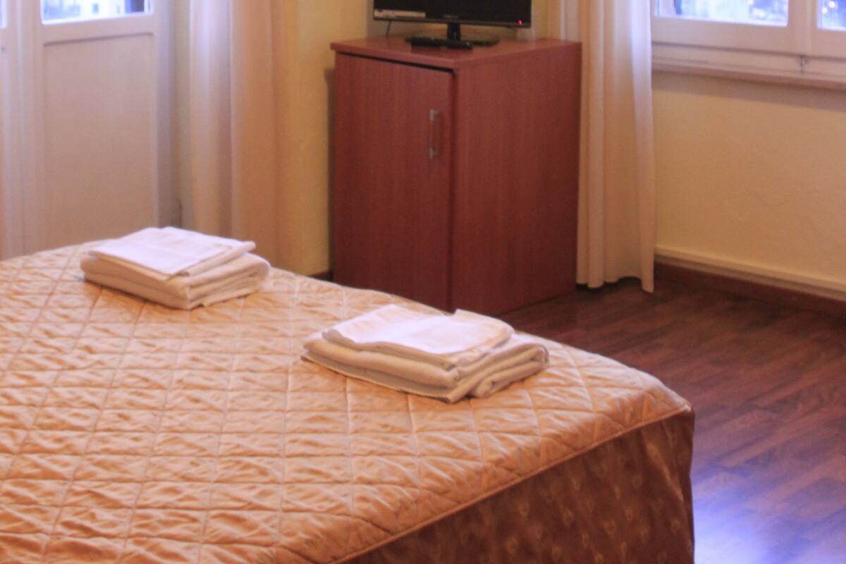 Une chambre d’hôtel avec un lit, un téléviseur posé sur une commode en bois et des rideaux aux fenêtres.